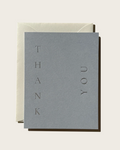 A minimalist thank you card in textured gray with the words 'THANK YOU' arranged vertically, shown with a cream envelope on a light beige background.