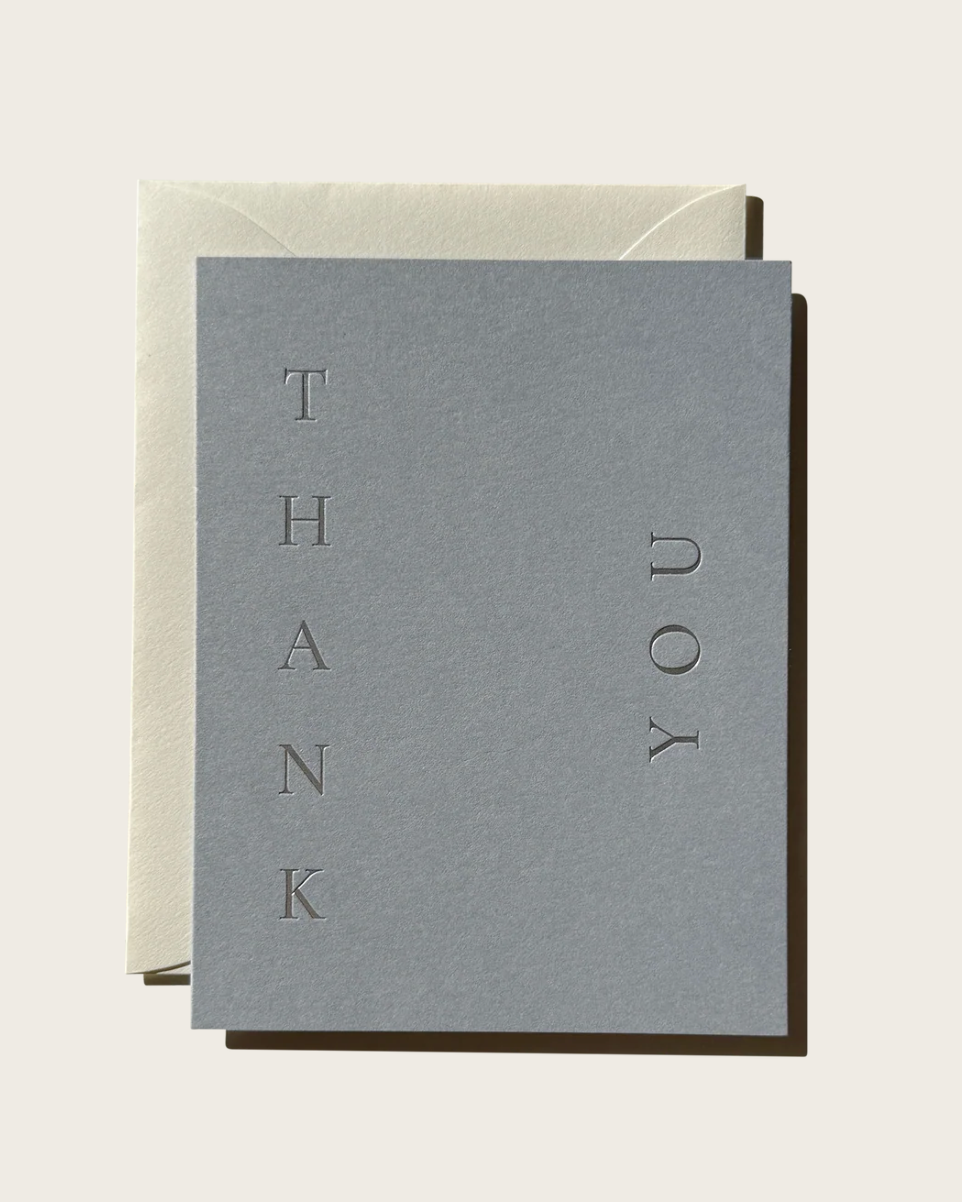 A minimalist thank you card in textured gray with the words 'THANK YOU' arranged vertically, shown with a cream envelope on a light beige background.