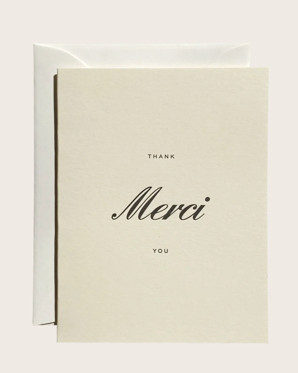 A minimalist thank you card in warm off-white with black text reading 'THANK Merci YOU' in elegant script and serif fonts, shown with a matching off-white envelope behind it on a light neutral background.